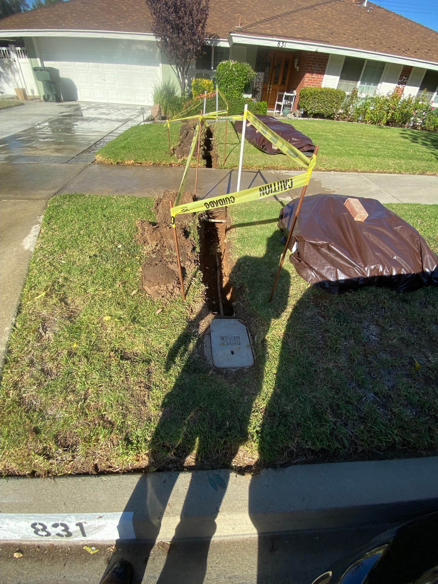 Trench running from the water meter box across a front yard toward the house, marked with caution tape ahead of city inspection